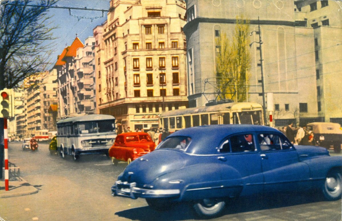 Bukarest, Nicolae Bălcescu sugárút, ötvenes évek vége. Ezen a fáintosan utószínezett képen néhány igazi különlegességet azonosíthatunk az akkor még igen gyér fővárosi forgalom résztvevőjeként. Akár egy vérszomjas kapitalista őslény, úgy uralja a szcénát a megkékített 1947-es Buick Roadmaster Mk4. Vajon hogy került ide, s vajon mi lett a sorsa? Talán épp egy vasfüggöny-leeresztés előtti hivatalos amerikai importból származik. A háttérben az utazni vágyó tömeg szelíden villamosozik, vagy TV-1-es busszal utazik, és többek között feltűnik egy GAZ-69-es terepjáró és egy pironkodó M-20-as Pobeda is.
